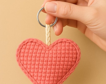 Peut inclure: Un porte-clés en forme de cœur crocheté de couleur corail. Le cœur est attaché à un porte-clés argenté avec une boucle tressée de couleur crème. Le fond est de couleur beige clair.