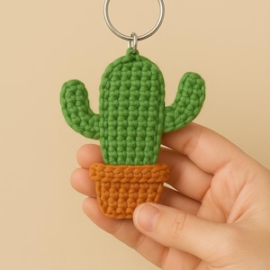 May include: A handmade crochet cactus keychain. The cactus is green with two arms, and sits in an orange pot. The keychain is attached to a silver metal ring. The item is held in a person's hand.