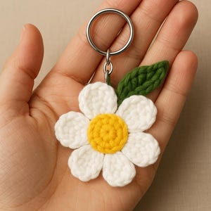 Może przedstawiać: Ręcznie robiony brelok do kluczy z kwiatem zrobionym na szydełku. Kwiat ma białe płatki i żółty środek, z zielonym liściem. Brelok jest przymocowany do srebrnego metalowego kółka.