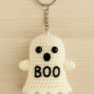 Puede incluir: Un llavero de fantasma de ganchillo blanco roto hecho a mano con ojos de bot&oacute;n negros y un dise&ntilde;o "BOO" negro. El fantasma tiene brazos peque&ntilde;os y est&aacute; unido a un llavero plateado con una cadena. El fondo es de vetas de madera clara.