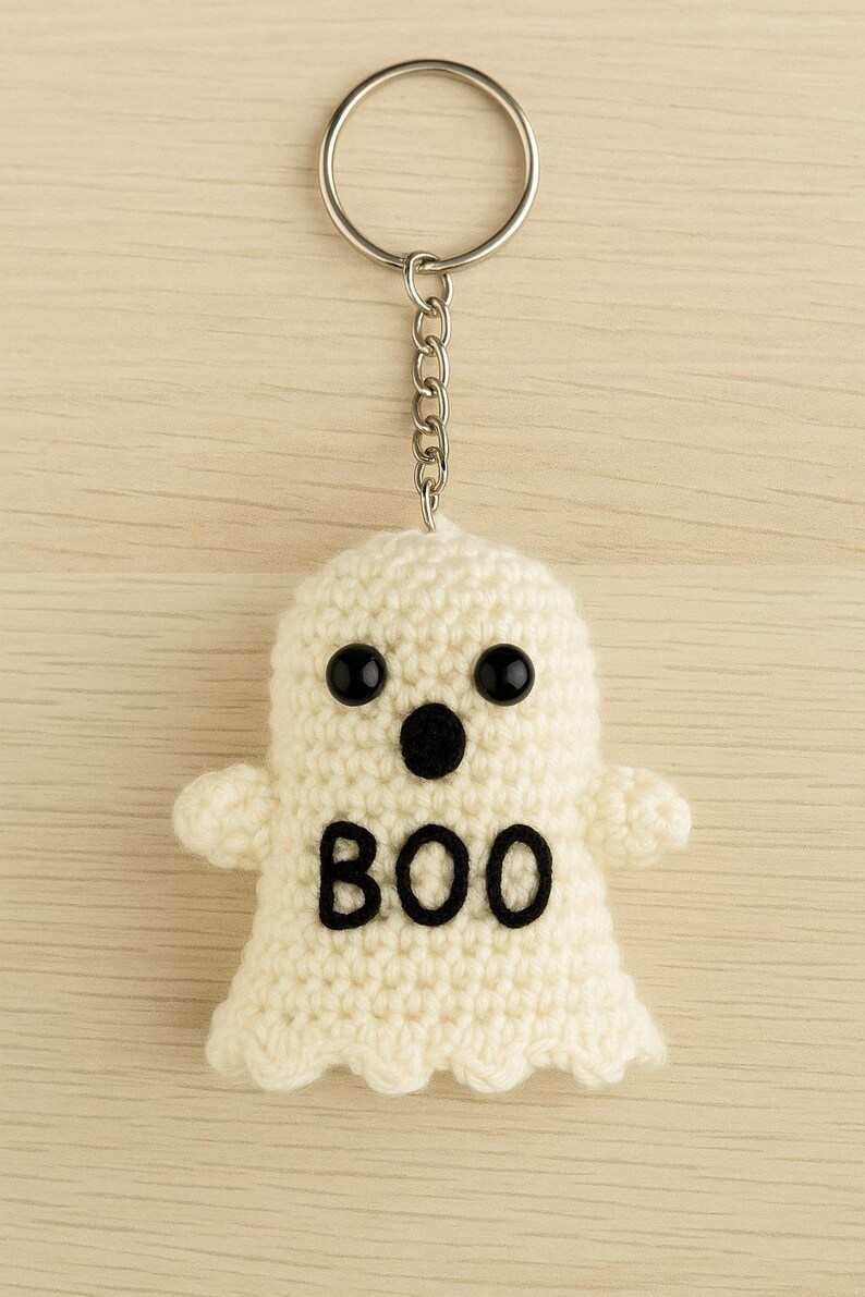 Puede incluir: Un llavero de fantasma de ganchillo blanco roto hecho a mano con ojos de bot&oacute;n negros y un dise&ntilde;o "BOO" negro. El fantasma tiene brazos peque&ntilde;os y est&aacute; unido a un llavero plateado con una cadena. El fondo es de vetas de madera clara.