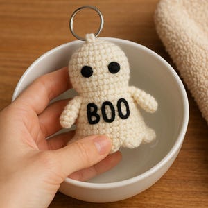 Puede incluir: Un llavero de fantasma de ganchillo color crema con ojos negros y la palabra "BOO" en negro. El fantasma tiene brazos peque&ntilde;os y un llavero plateado. El llavero est&aacute; en un cuenco blanco.