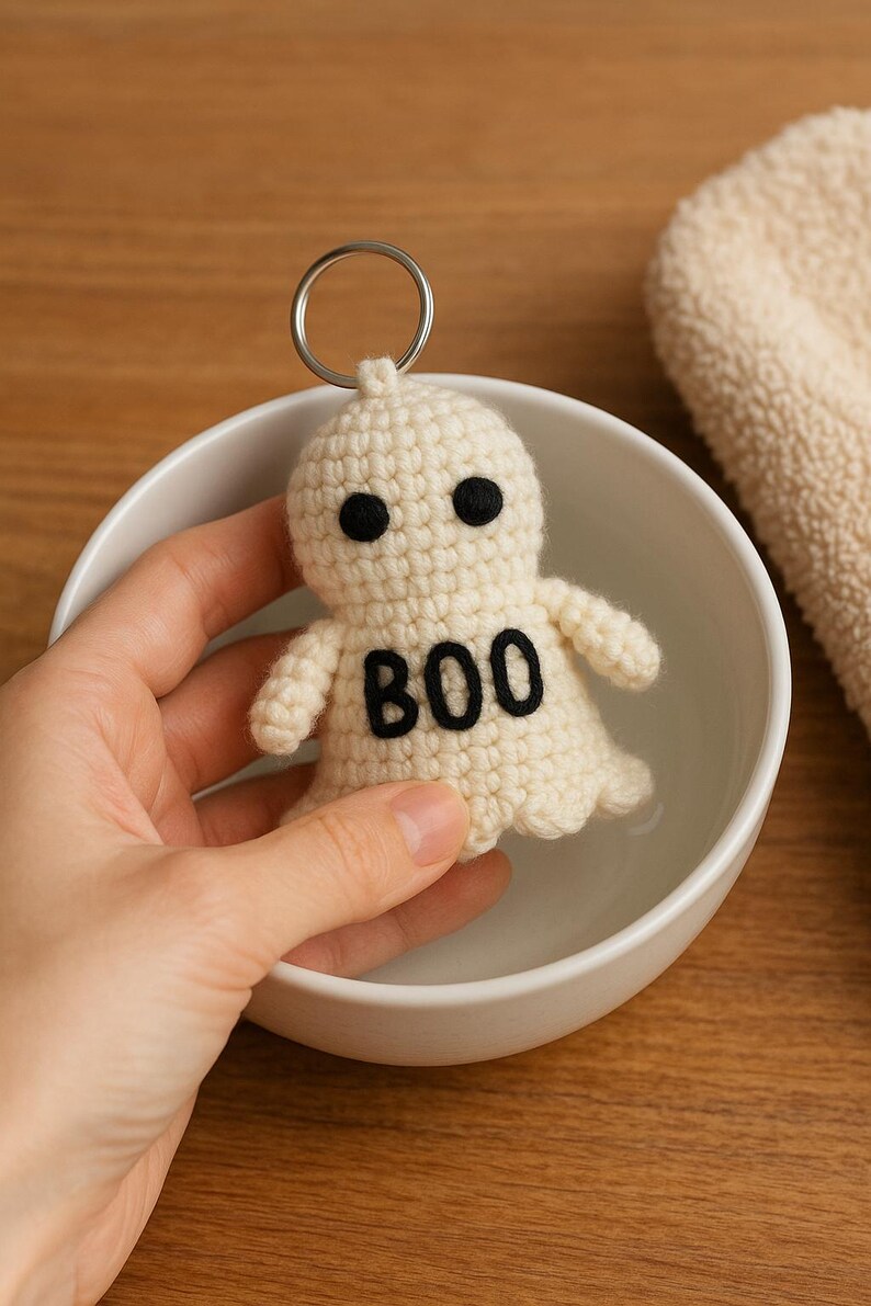 Puede incluir: Un llavero de fantasma de ganchillo color crema con ojos negros y la palabra "BOO" en negro. El fantasma tiene brazos peque&ntilde;os y un llavero plateado. El llavero est&aacute; en un cuenco blanco.