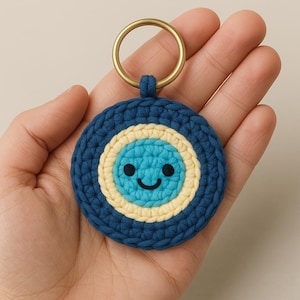 May include: A handmade crochet keychain featuring a smiling face design. The keychain has a dark blue outer ring, a light yellow middle ring, and a light blue center with a smiling face. It includes a gold-colored metal ring for attaching keys.
