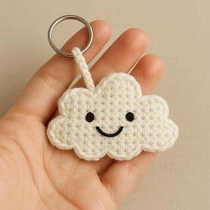 Könnte beinhalten: Ein handgefertigter, cremefarbener Häkel-Wolken-Schlüsselanhänger mit einem lächelnden Gesicht. Die Wolke hat schwarze Augen und einen gebogenen Mund. Ein silberner Metall-Schlüsselring ist oben an der Wolke befestigt.