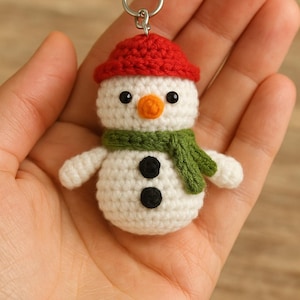 Może przedstawiać: Szydełkowy brelok do kluczy w kształcie bałwana z czerwonym kapeluszem, zielonym szalikiem i czarnymi guzikami. Bałwanek ma białe ciało, pomarańczowy nos i czarne oczy. Srebrny brelok jest przymocowany.