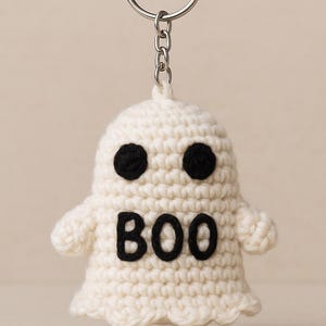 Puede incluir: Llavero de fantasma de ganchillo blanco con ojos negros y la palabra "BOO" en negro. El fantasma tiene brazos peque&ntilde;os y un llavero plateado adjunto en la parte superior.