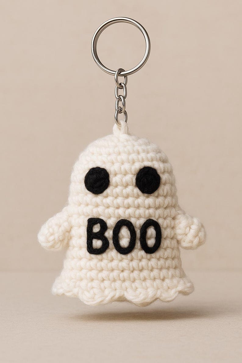 Puede incluir: Llavero de fantasma de ganchillo blanco con ojos negros y la palabra "BOO" en negro. El fantasma tiene brazos peque&ntilde;os y un llavero plateado adjunto en la parte superior.