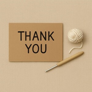 Op de afbeelding: Een kraftpapieren kaart met de zwarte tekst "THANK YOU". Een bolletje witte wol en een houten haaknaald liggen naast de kaart. De achtergrond is een beige stof.