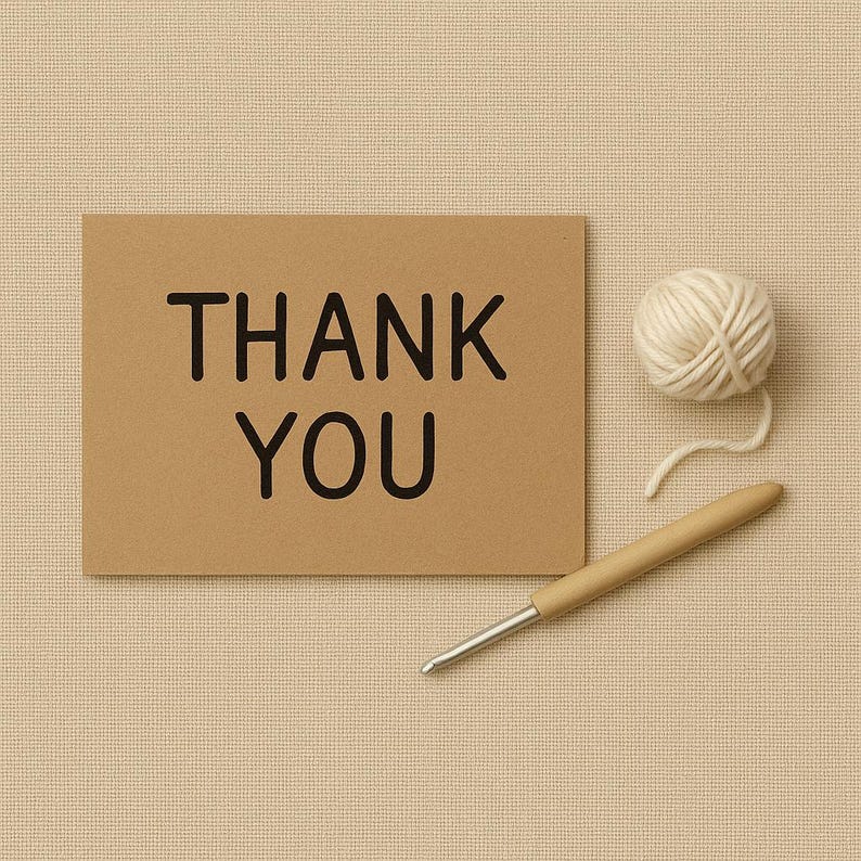 Op de afbeelding: Een kraftpapieren kaart met de zwarte tekst "THANK YOU". Een bolletje witte wol en een houten haaknaald liggen naast de kaart. De achtergrond is een beige stof.