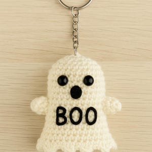 Könnte beinhalten: Ein handgefertigter, cremefarbener Geister-Schlüsselanhänger aus Häkelarbeit mit schwarzen Knopfaugen und einem schwarzen "BOO"-Design. Der Geist hat kleine Arme und ist an einem silberfarbenen Schlüsselring mit einer Kette befestigt. Der Hintergrund ist eine helle Holzmaserung.