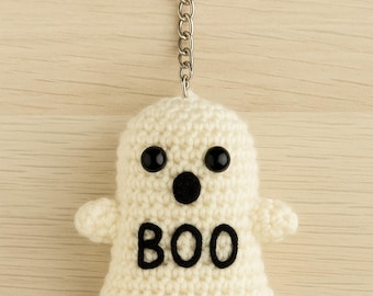 Puede incluir: Un llavero de fantasma de ganchillo blanco roto hecho a mano con ojos de botón negros y un diseño "BOO" negro. El fantasma tiene brazos pequeños y está unido a un llavero plateado con una cadena. El fondo es de vetas de madera clara.