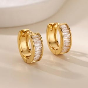 Peut inclure: Une paire de créoles dorées ornées d'une rangée de pierres transparentes rectangulaires taillées en baguette sur la face avant. Ces boucles d'oreilles présentent un mécanisme de fermeture à charnière et une finition métallique polie.