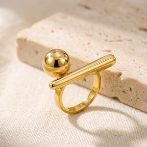 Peut inclure: Une bague dorée composée d'une perle sphérique polie fixée sur une barre cylindrique horizontale. L'anneau est circulaire et lisse, se connectant à la base de la barre. L'ensemble présente une finition métallique réfléchissante et épurée.