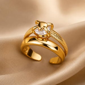 Peut inclure: Une bague ouverte dorée ornée d'une pierre transparente taille ronde, maintenue par un sertissage en forme de cœur. L'anneau se compose de deux parties distinctes : une section lisse et polie et une section texturée à motifs croisés.