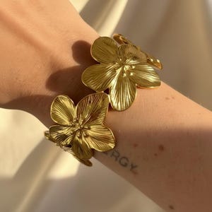 Peut inclure: Un bracelet doré composé d'une série de breloques en forme de fleurs à cinq pétales reliées entre elles. Chaque fleur présente une surface texturée avec des motifs rayonnants et un centre en relief. Le métal a une finition polie et brillante.