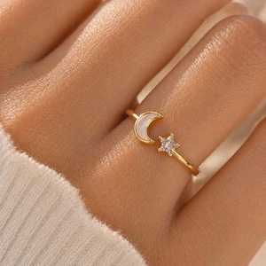 Peut inclure: Une bague dorée ornée d'un croissant de lune et d'une étoile. La lune présente un incrustation nacrée blanche, et l'étoile est ornée d'une pierre précieuse transparente. La bague est portée au doigt.