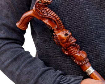 May include: A wooden walking cane with a carved cobra head and a skull design. The cane is brown with a silver band near the bottom. The cobra has green eyes.