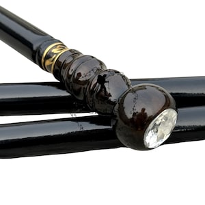 Könnte beinhalten: Schwarzer Holz-Gehstock mit dekorativem Griff. Der Griff ist dunkelbraun, abgerundet und hat am Ende einen klaren, facettierten Kristall. Ein goldfarbenes Band akzentuiert die Oberseite des Griffs.