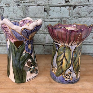 May include: Two ceramic candle holders with scalloped rims, painted with purple and blue floral designs, green leaves, and small bee illustrations. Each piece features cut-out sections and the signature Heather Goldminc on the lower base.