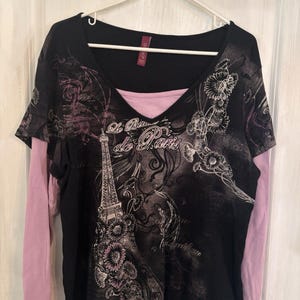May include: A black long-sleeve shirt with a layered look featuring light purple sleeves and a matching inner neckline. The front displays a graphic print of the Eiffel Tower, floral motifs, and the text "Le Beau de Paris" in white script.