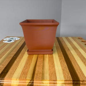 May include: A square, terracotta-colored planter with a matching base. The planter has a slightly flared design and sits on a wooden surface with alternating light and dark wood grain stripes. A sticker is visible on the left.