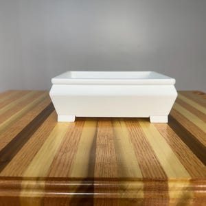 May include: A white, rectangular planter box with a raised rim and small feet. The planter is made of a solid material and sits on a wooden table with alternating light and dark wood stripes.