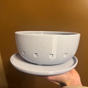 May include: A light blue speckled ceramic planter with a matching saucer. The bowl-shaped planter has square cutouts around the rim. The planter and saucer are designed for indoor or outdoor use.