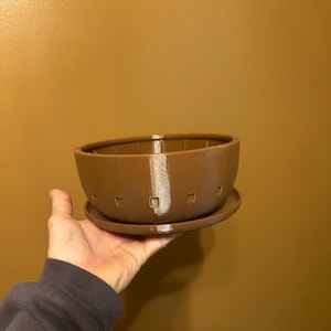 May include: A brown, 3D-printed planter with a matching saucer. The planter has a bowl shape with a series of square cutouts around the rim and a vertical white stripe. The saucer is round and flat.