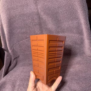 May include: A brown, rectangular planter with a geometric design. The planter has a series of rectangular and horizontal line patterns. The planter is held by a hand against a purple background.