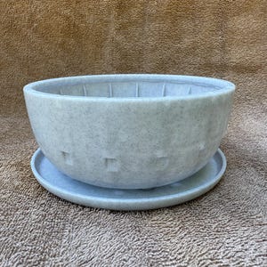 Puede incluir: Una maceta de cerámica moteada azul claro con un platillo a juego. La maceta en forma de cuenco tiene una superficie texturizada y cortes cuadrados. La maceta está diseñada para uso en interiores o exteriores y es adecuada para varias plantas.