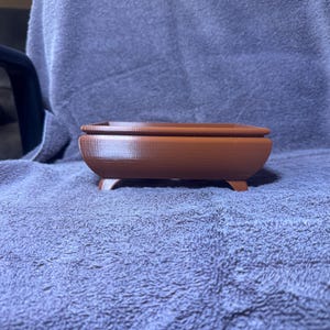 Puede incluir: Un recipiente rectangular marrón con tapa, apoyado sobre cuatro patas pequeñas. El recipiente tiene un diseño liso y curvado y está hecho de un material sólido. El recipiente está sobre una superficie azul-gris texturizada.