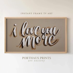 May include: A framed art print featuring the phrase "i love you MORE" in white script with brown shading. The artwork is set against a tan backdrop and enclosed within a light wood frame. The text "INSTANT FRAME TV ART" and "PORTHAUS PRINTS art society" are also visible.