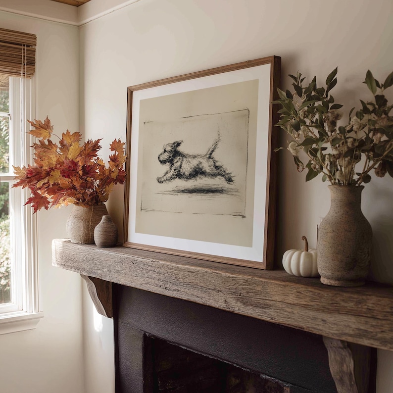 May include: A framed print of a running dog in a living room setting. The artwork is in a light wood frame and is displayed above a dark wooden mantel. Decorative vases and fall foliage are arranged on the mantel.