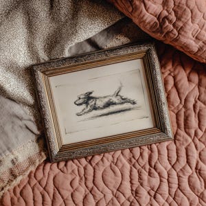 May include: A framed pencil sketch of a dog in mid-leap. The artwork is in a gold-colored frame with a detailed border. The dog is depicted in motion, with its legs extended. The background is a neutral color.