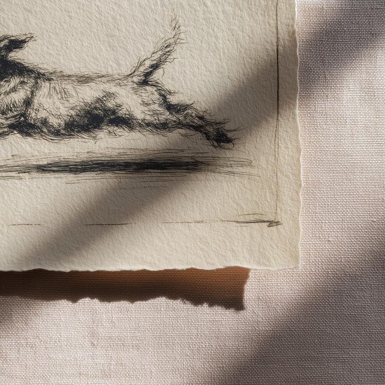 May include: A black and white etching of a dog in mid-stride, with a textured, detailed appearance. The artwork is on a piece of paper with a torn edge, set against a neutral fabric background. The dog's fur is meticulously rendered.