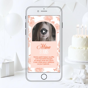 Peut inclure: Un smartphone affiche une vidéo d'anniversaire avec une photo d'enfant et le texte "Mina 3 ANS". L'écran est encadré d'un motif floral. Un gâteau blanc avec des bougies et des ballons en arrière-plan, suggérant une fête.