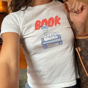 以下が含まれることがあります： 眼鏡をかけた漫画の虫が本の上に乗り、その上に赤い文字で「BOOK」と書かれたグラフィックがプリントされた白い半袖Tシャツ。Tシャツは青いデニムジーンズと茶色の柄入りショルダーバッグと組み合わされています。