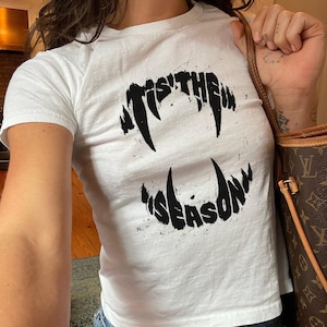 May include: A white short-sleeved t-shirt featuring a black graphic print of sharp, jagged vampire fangs arranged in a circular shape. The text "'TIS THE" is printed along the top set of fangs, and "SEASON" is printed along the bottom set of fangs.