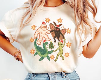 May include: An ivory short-sleeve t-shirt featuring a graphic print of a white yeti, a black mothman, a green dinosaur, and a green alien, all wearing red and white Santa hats. The characters are dancing around a small Christmas tree with stars.