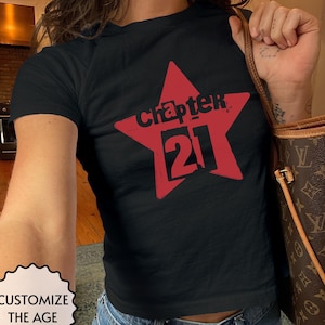 May include: A black short-sleeved t-shirt featuring a large red star graphic on the chest. Inside the star, the word "chapter" is printed in black above the large black number "21". A small white badge in the corner reads "CUSTOMIZE THE AGE".