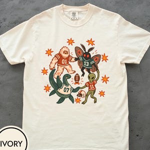 May include: An ivory short-sleeve t-shirt featuring a graphic print of a Bigfoot, a moth creature, a sea monster, and an alien wearing football jerseys numbered 01, 13, 07, and 11. The design includes a football, a helmet, and orange star shapes.