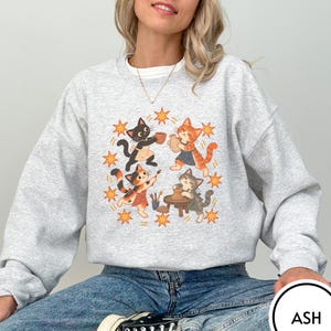 May include: An ash gray crewneck sweatshirt featuring a graphic print of four cats wearing aprons and working with pottery. The cats are surrounded by orange star shapes. The sweatshirt has long sleeves and a ribbed collar, cuffs, and hem.