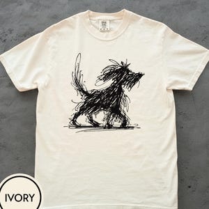 May include: Ivory-coloured t-shirt with a black, scribbled illustration of a dog. The dog is shown walking, with a long tail and shaggy fur. The shirt has a crew neck and short sleeves. The word "IVORY" is in a circle at the bottom left.
