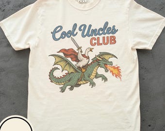 May include: An ivory short-sleeve t-shirt featuring the text Cool Uncles CLUB in blue and red lettering. The graphic depicts a white goose wearing a red cape and holding a sword while riding a green dragon that is breathing orange and yellow fire.
