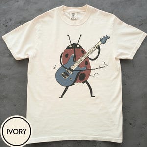Puede incluir: Una camiseta de manga corta color marfil con una ilustración gráfica de una mariquita roja con puntos negros tocando una guitarra eléctrica azul. La mariquita tiene antenas y extremidades negras finas, el diseño está centrado en el frente.