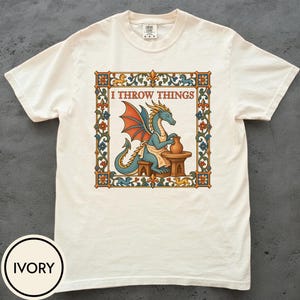 May include: An ivory short-sleeve t-shirt featuring a graphic of a blue dragon wearing an apron and working at a pottery wheel. The design is enclosed in a decorative floral border with the text I THROW THINGS printed in brown above the dragon.