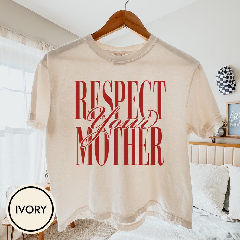 May include: An ivory short-sleeve t-shirt featuring the red text "RESPECT YOUR MOTHER" printed on the front. The shirt has a crew neckline and is displayed on a wooden hanger. The text is arranged with "RESPECT" and "MOTHER" in a tall, bold font, while "YOUR" is written in a cursive script.