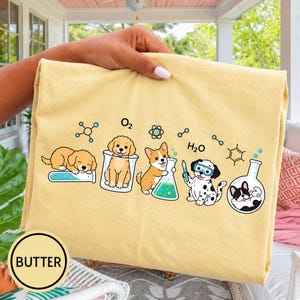 Könnte beinhalten: Ein blassgelbes T-Shirt mit einem Grafikdruck von fünf Cartoon-Welpen, die mit Laborgeräten und chemischen Symbolen interagieren. Das Design zeigt einen Golden Retriever, einen Pudel, einen Corgi, einen Dalmatiner und eine Französische Bulldogge.