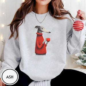 May include: Light gray sweatshirt featuring a whimsical illustration of a greyhound dog wearing a red sweater and holding a red mug. The dog is depicted in a cartoon style. The sweatshirt has a crew neck and long sleeves. The word "ASH" is in a white circle.